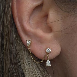 May include: A gold double-stud earring connected by a delicate chain. One stud features a pear-shaped stone, while the other stud is round with a dangling pear-shaped stone. The jewelry is crafted from polished yellow gold and clear gemstones.