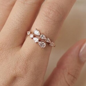 Op de afbeelding: Een roségouden ring met een bypass-ontwerp en meerdere peervormige, heldere stenen langs de band. De ring heeft een gepolijste metalen afwerking en de stenen zijn zo gezet dat ze elkaar op de vinger enigszins overlappen.