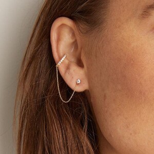 May include: A gold ear cuff with a chain connecting to a small stud earring. The ear cuff is adorned with small, sparkling stones. The jewelry is worn on the ear of a person wearing a white shirt.