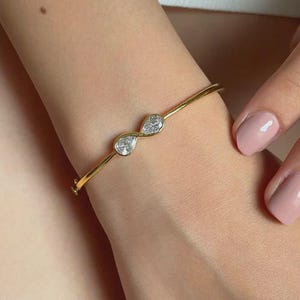 May include: A gold bangle bracelet featuring two pear-shaped, clear gemstones. The bracelet is worn on a wrist, showcasing the delicate design and the sparkle of the gemstones. The bracelet has a simple, elegant design.
