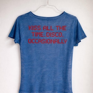 May include: Blue t-shirt with the text "KISS ALL THE TIME. DISCO, OCCASIONALLY" in red. The shirt is hanging on a wire hanger against a white background. The t-shirt has short sleeves and a scoop neck.