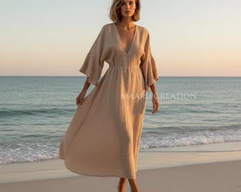 May include: A beige, flowing dress with a deep V-neck and wide sleeves. The dress is cinched at the waist and falls to the ankles. The text "MAARU CREATION" is visible on the dress. The background shows a beach and ocean.