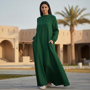May include: A long, emerald green dress with long sleeves and pockets. The dress has a high neckline and a flowing silhouette. The model is wearing embellished flats. The background features a building with arches and a palm tree.