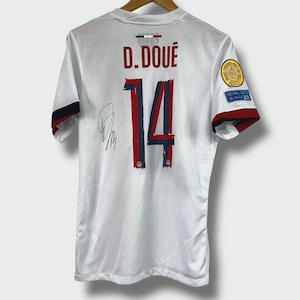 Pode incluir: Camisola de futebol branca com o nome "D. DOUÉ" e o número "14" em vermelho e azul. A camisola tem uma assinatura no lado esquerdo e um patch na manga direita. As mangas têm acabamentos vermelhos e pretos.