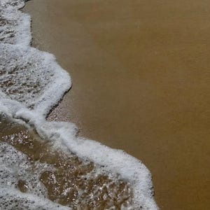 Może przedstawiać: Zdjęcie sceny na plaży. Obraz przedstawia spotkanie oceanu i piasku. Białe, spienione fale delikatnie toczą się na brzeg, kontrastując z gładkim, jasnobrązowym piaskiem. Scena wywołuje poczucie spokoju i naturalnego piękna.