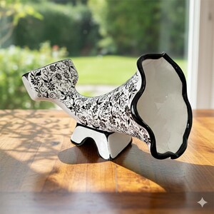 May include: A white ceramic wine bottle holder with a black floral pattern. The holder has a curved shape with a wide opening and a black rim. It rests on a white base with black accents, set on a wooden surface.