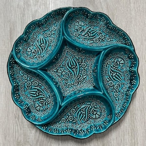 Puede incluir: Una fuente de cerámica turquesa con cinco compartimentos. La fuente presenta un intrincado diseño floral y de cachemira, con un borde festoneado. El color es un verde azulado vibrante, y la fuente parece estar esmaltada.