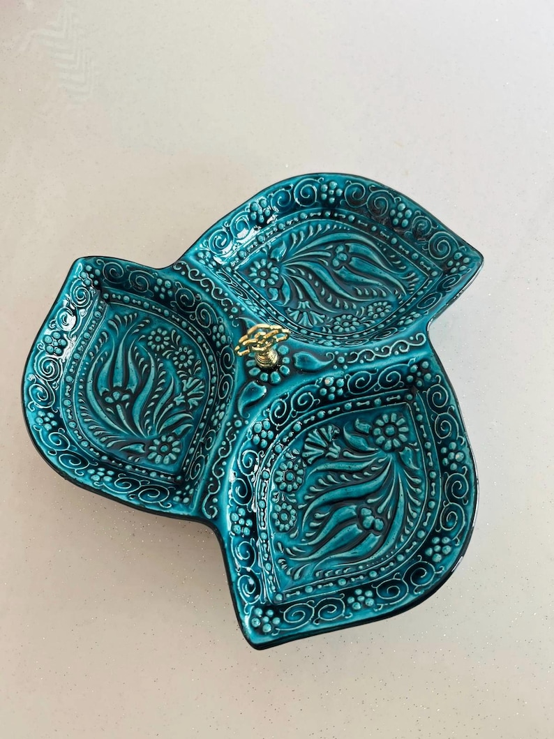Puede incluir: Una fuente de cer&aacute;mica turquesa con tres secciones, con intrincados patrones florales y de remolinos. La fuente tiene un asa dorada en el centro. El dise&ntilde;o est&aacute; en relieve, creando una superficie texturizada. La fuente se utiliza probablemente para servir aperitivos.