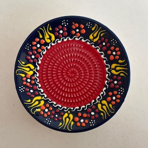May include: A round, decorative plate with a dark blue rim and a red center. The rim features yellow, orange, and white painted designs. The center has a textured, grater-like surface.