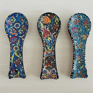 Puede incluir: Tres reposacucharas de cerámica con intrincados diseños florales. Cada uno presenta un patrón único de flores rojas, naranjas, amarillas y azules sobre un fondo azul oscuro. Los bordes están delineados con puntos blancos, creando un accesorio de cocina decorativo.