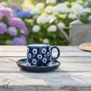 May include: A blue ceramic coffee cup and saucer set with a white floral pattern. A small silver spoon rests on the saucer. The cup and saucer are on a weathered wooden table, with a straw hat and garden in the background.