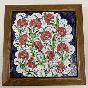 May include: A framed ceramic tile featuring a floral design. The tile has a dark blue background with a white scalloped border. Red flowers with green leaves and blue accents are arranged across the tile. The frame is made of brown wood.