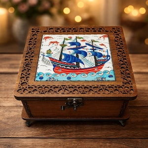 May include: A wooden jewelry box with a decorative carved frame. The lid features a colorful illustration of a sailboat on a blue sea. The box is brown with a metal clasp and rests on a wooden surface.