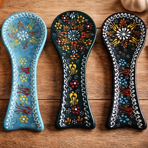 Puede incluir: Tres reposacucharas de cerámica con intrincados diseños florales. Los reposacucharas son en tonos azul, verde y negro, con acentos coloridos. Están sobre una superficie de madera, con ajo y especias en el fondo.