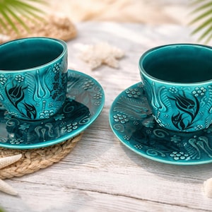 May include: Two turquoise ceramic teacups and saucers with a floral design. The cups and saucers are placed on a light-colored wooden surface, with a starfish, seashells, and lemon slice nearby. The cups have handles.