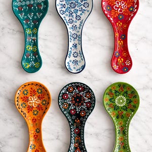 Könnte beinhalten: Sechs farbenfrohe Keramik-Löffelablagen mit aufwendigen Blumenmustern. Die Löffelablagen sind in den Farben Türkis, Weiß, Rot, Orange, Schwarz und Grün gehalten. Jede Löffelablage hat ein einzigartiges Design mit einem gebogenen Griff und einer ovalen Schale.