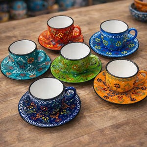 Könnte beinhalten: Sechs farbenfrohe Keramik-Kaffeetassen und Untertassen. Jede Tasse und Untertasse hat ein einzigartiges Blumenmuster in Rot-, Blau-, Grün- und Orangetönen. Die Tassen haben weiße Innenräume und stehen auf einer Holzoberfläche.