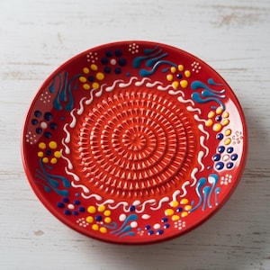 May include: A red ceramic plate with a textured center and decorative blue, white, and yellow accents. The plate has a circular design with a spiral pattern in the center.