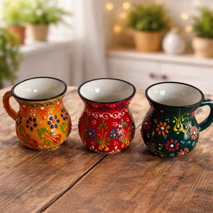 May include: Three colorful ceramic mugs with floral patterns. The mugs are orange, red, and green, each with a white interior and a black rim. They sit on a wooden surface, showcasing their unique designs and shapes.