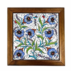 May include: Square decorative tile with a floral design. The tile features blue flowers with black centers, green leaves, and red accents on a white background. The tile is framed in a dark brown wooden frame.