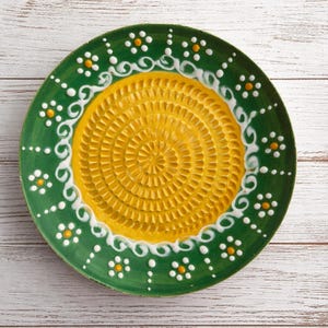 Könnte beinhalten: Ein runder Keramik-Reibeteller mit lebendigem Design. Die Mitte ist gelb mit einer strukturierten Oberfläche zum Reiben. Der Rand ist grün, verziert mit weißen Wirbeln und kleinen gelb-weißen floralen Akzenten. Ein dekorativer Küchenartikel.