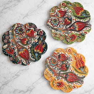 May include: Three decorative coasters with a floral design. Each coaster has a unique colour scheme: dark blue, green, and yellow. The floral pattern includes red, orange, and blue accents. The coasters are shaped like a stylized flower.