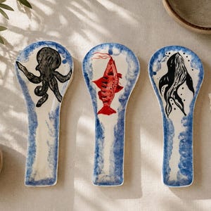 Pode incluir: Três apoios de colher em cerâmica com base cor de creme e bordas pintadas de azul. Cada peça apresenta uma criatura marinha pintada à mão: um polvo preto, um peixe vermelho e uma baleia preta, centrados na forma oval de cada suporte.