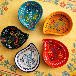 Puede incluir: Cinco cuencos de cerámica coloridos en forma de lágrima con intrincados diseños florales. Los cuencos son de color azul, naranja, rojo, negro y blanco, cada uno con diseños únicos. Se encuentran sobre una superficie amarilla con un patrón floral.