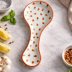 Puede incluir: Un reposacucharas de cerámica blanca con borde naranja, decorado con pequeños motivos de frutas naranjas y tallos verdes. La imagen incluye ingredientes como albahaca, ajo y limones, sugiriendo un tema culinario.