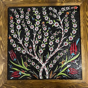 May include: Square ceramic tile with a black background, featuring a white tree design with white flowers and red centers. The tile is framed in a light brown wooden frame. Decorative art.