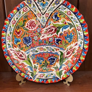 May include: A decorative ceramic plate with a vibrant floral design. The plate features a red, orange, and white color scheme with blue and green accents. The intricate pattern includes flowers and leaves. The plate is displayed on a wooden stand.