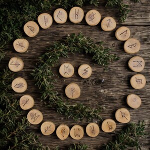 Peut inclure: Ensemble de pierres de runes en bois disposées en cercle sur une surface en bois rustique, entourées de brins verts. Chaque pierre circulaire présente un symbole gravé unique en noir, créant une esthétique mystique et naturelle.