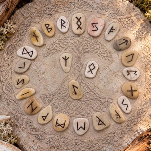 Puede incluir: Una losa de piedra circular grabada con un diseño de árbol sirve de base para veinticinco piedras lisas y claras. Cada piedra presenta un símbolo rúnico negro único pintado en su superficie, dispuesto en un patrón circular.
