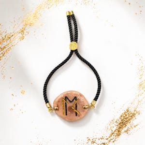 May include: A bracelet featuring a light brown, oval-shaped charm with gold-coloured accents and black rope. The charm has gold and black markings. The bracelet has a gold-coloured clasp and a gold-coloured leaf design.