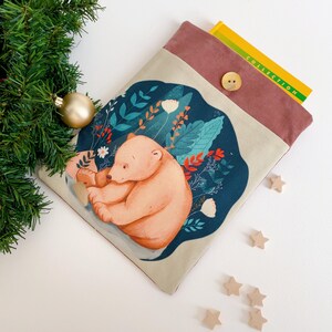 May include: A beige and burgundy book sleeve featuring a watercolor illustration of a bear nestled in a floral design. The sleeve has a button closure and holds a yellow book. A gold Christmas ornament and wooden stars are in the background.