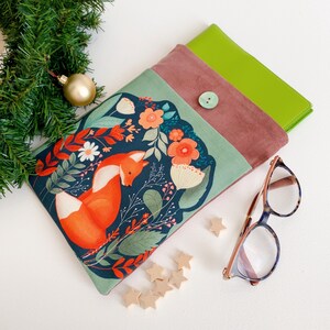 May include: A fabric book sleeve featuring a fox illustration with orange flowers and green leaves on a teal background. The sleeve has a button closure and a green interior. A pair of glasses and wooden stars are next to the sleeve.