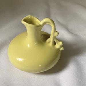 May include: A small, pale yellow ceramic pitcher with a rounded body and a curved handle. The pitcher has a spout and a decorative element on the handle. The overall design is simple and elegant, suitable for a variety of uses.