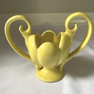 May include: A pale yellow ceramic vase with a tulip-like design. The vase has two curved handles and a scalloped rim. The base is round and the overall shape is symmetrical. The vase is likely vintage and would be a decorative item.