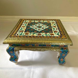 May include: A decorative, square table with a gold-colored frame and legs. The tabletop features a colorful floral pattern in shades of green, orange, and white. The legs are adorned with blue and floral designs. The table is approximately 12 inches long and wide.