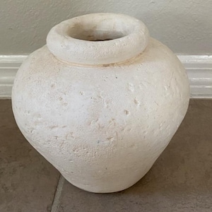 May include: A textured, off-white ceramic vase with a rounded body and a cylindrical neck. The vase has a rough, pitted surface and a wide opening. The neutral color and simple design make it suitable for various decor styles.