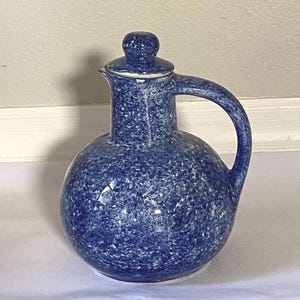 May include: A blue ceramic pitcher with a speckled pattern. The pitcher has a round body, a curved handle, and a lid with a small knob. The color is a deep blue with white speckles, giving it a rustic appearance.