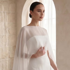 May include: A floor-length white gown featuring a strapless bodice and a sheer, lightweight cape overlay that drapes over the shoulders and arms. The garment is made of solid white fabric with a smooth texture and a translucent, flowing chiffon layer.