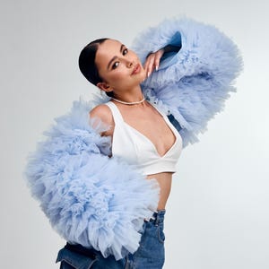 Puede incluir: Chaqueta de tul azul claro con mangas voluminosas. La chaqueta se combina con un top corto blanco y vaqueros. Un collar de perlas añade un toque de elegancia. El conjunto se presenta sobre un fondo blanco.