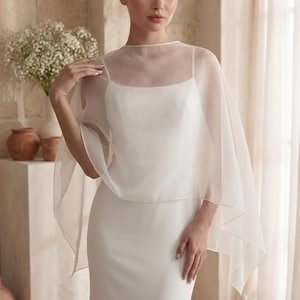 May include: A sheer white organza capelet worn over a simple white spaghetti strap dress. The capelet features a high round neckline and drapes loosely over the shoulders, creating a soft, layered look with a clean, minimalist aesthetic.