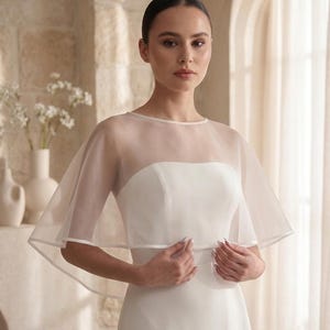May include: A sheer white organza capelet featuring a round neckline and wide, flowing short sleeves. The capelet is worn over a strapless white gown, with a clean, minimalist design and a delicate hemline that drapes gracefully over the shoulders.