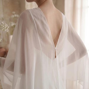 May include: A sheer white garment featuring a deep V-neckline that extends down the back. The piece has wide, flowing sleeves and a lightweight, semi-transparent fabric construction that drapes loosely over the shoulders and upper torso area.