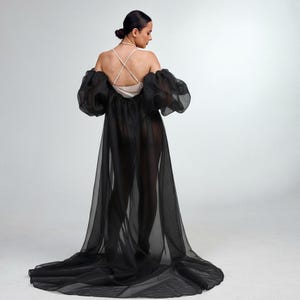 May include: A black sheer robe with large, puffed sleeves and a long train. The robe is worn over a cream-coloured top with thin straps. The model is seen from the back, with hair in a bun, against a white background.