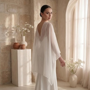 May include: A floor-length white gown featuring a sheer, flowing cape overlay with a deep V-shaped back. The garment is constructed from a solid opaque fabric base with a lightweight, translucent chiffon-style material draped over the shoulders.