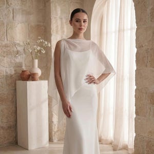 May include: A floor-length white gown featuring a sleeveless bodice with a scoop neckline and a sheer, lightweight chiffon overlay that drapes over the shoulders and arms, creating a soft, flowing silhouette with a clean, solid white finish.