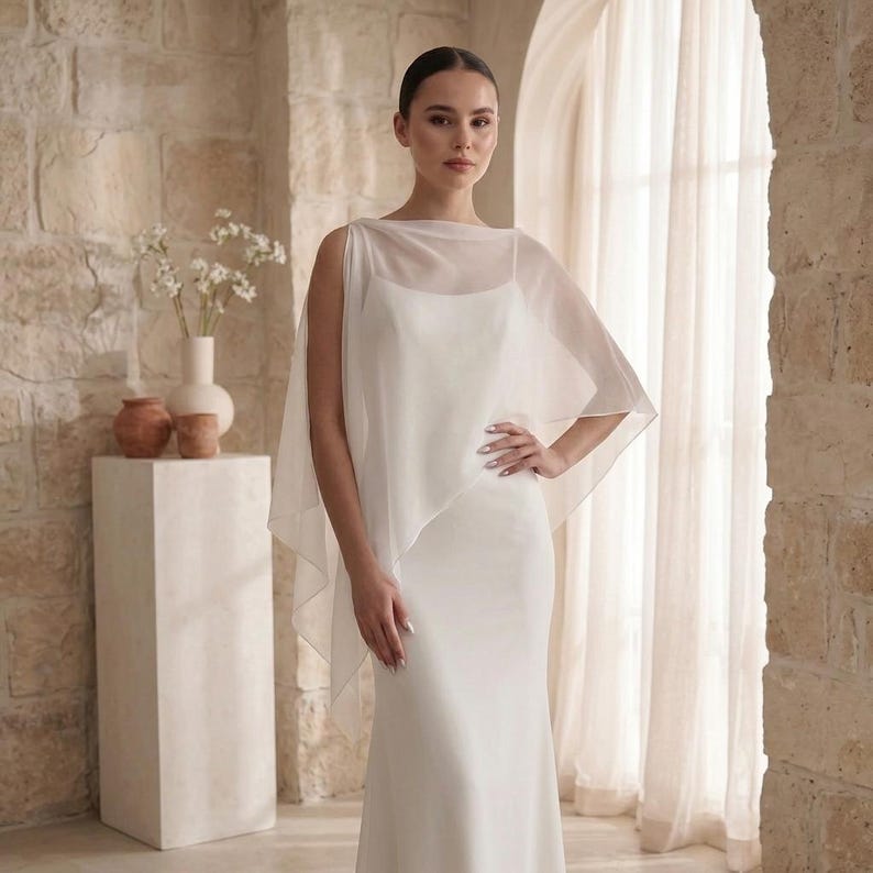 May include: A floor-length white gown featuring a sleeveless bodice with a scoop neckline and a sheer, lightweight chiffon overlay that drapes over the shoulders and arms, creating a soft, flowing silhouette with a clean, solid white finish.